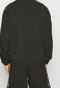 Chaqueta de atletismo negra con cintura y puños elásticos, que presenta una textura suave y un diseño sutil en las mangas.