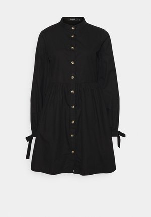 Sort langærmet kjole med højt krave, forgyldte knapper foran, indsnævret talje og bindedetaljer ved manchetterne.