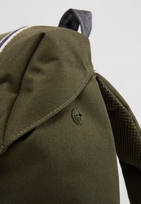 Olivengrøn rygsæk med et struktureret lærredsmateriale, flaplukning og mesh-detaljer på siden. Har en rund logoknap.