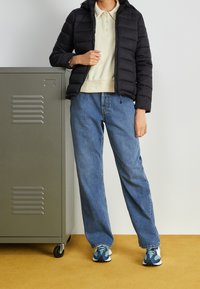 Czarna kurtka puchowa z kapturem, beżowa koszulka polo, jasnoniebieskie luźne dżinsy i kolorowe sneakersy. W tle znajduje się szary schowek.