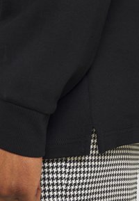 Mustat collegepaita, jossa on ribbiteksturointi ja halkio helmoissa, päällä mustavalkoinen houndstooth-kuvioitu vaate. Lähikuva.