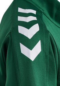 Chaqueta deportiva verde con una textura suave, que presenta logotipos en forma de chevrón blancos en el hombro izquierdo y un acabado de costura limpio.