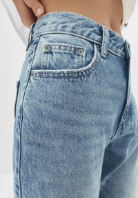 Jean en denim bleu clair avec une coupe ajustée, cinq poches et des détails de couture visibles le long de la taille et des coutures.