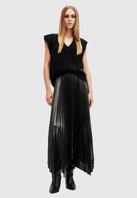 Zwart bontvest met een V-hals, gecombineerd met een lange geplooide zwarte rok. De rok heeft een glanzende textuur en een asymmetrische zoom. Zwarte enkellaarsjes.