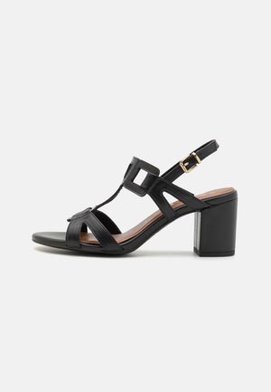 Sort hæl sandals med blokhæl, der har et T-strap design, åben tå og justerbar ankelrem med guld spænde accenter.
