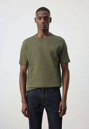 Jeune homme debout portant une chemise olive à manches courtes et un jean bleu foncé sur un fond clair uni.