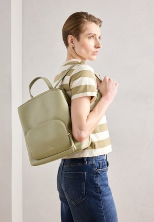 Personne aux cheveux courts portant une chemise rayée et un jean, porte un sac à dos vert clair sur une épaule, regardant sur le côté.