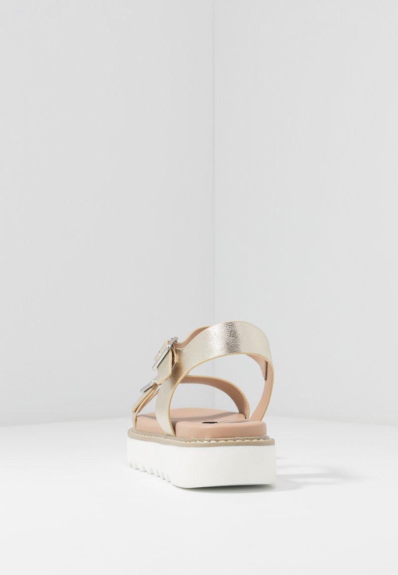 Sandale métallique dorée avec une bride ajustable, semelle intérieure beige douce et semelle blanche épaisse avec des rainures texturées pour plus de traction.