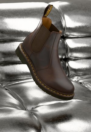 Brown leather Chelsea boot with mesh side panels, featuring a yellow stitched sole and a pull tab at the heel, resting on a metallic surface.