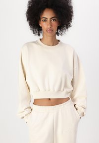 Lys beige avkortet hettegenser med lange ermer og ribbestrikkede mansjetter, med et preget logo på brystet, kombinert med matchende bukser med snøring.