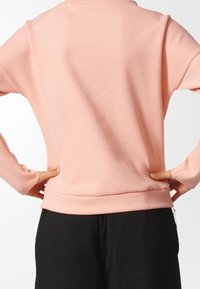 Persona con una camisa de manga larga rosa claro, con las manos en las caderas y pantalones negros con textura, visto desde atrás.