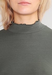 Dark green ribbed top with a high, ruffled neckline. The fabric has a soft texture and a form-fitting cut, suitable for layering.