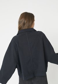 Marineblaue, taillierte Jacke mit weißen vertikalen Streifen, ausgestattet mit einem spitzen Kragen und weiten Ärmeln, hergestellt aus einem glatten, baumwollähnlichen Stoff.