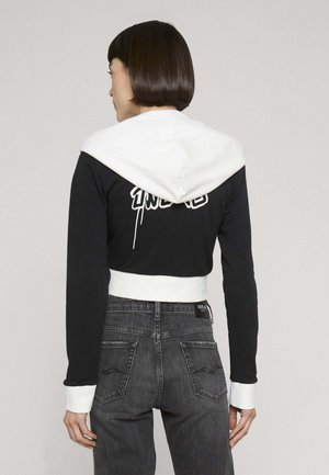 Femme aux cheveux courts et foncés portant un sweat à capuche court noir avec capuche et poignets blancs, et un jean noir délavé taille haute, vue de dos.