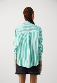 Grün und weiß gestreiftes Hemd mit einem Rückenfalten, lange Ärmel und einen spitzen Kragen. Getragen über einem schwarzen Jeansrock.