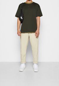 Camiseta de manga corta verde oscuro con puños y bolsillo beige, combinada con pantalones tapered de color beige claro con bolsillos laterales y una riñonera negra.