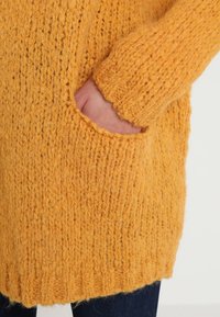 Knitted cardigan in bright yellow, showcasing thick, textured wool with a side pocket and ribbed hem.