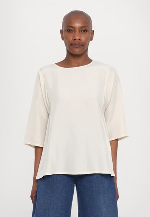 Blusa di colore crema, leggera, con scollatura tonda e maniche 3/4. Tessuto morbido con un fit rilassato, abbinata a jeans larghi blu.