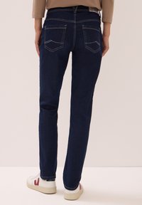 Persona vistiendo jeans de mezclilla azul oscuro y zapatillas blancas con detalles rojos, de pie frente a un fondo claro y liso.
