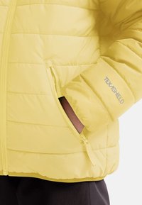 Chaqueta acolchada amarilla con diseño de cuadrícula, bolsillo lateral con cremallera y logo "TEXASHIELD" en la manga. Textura suave y ligera.