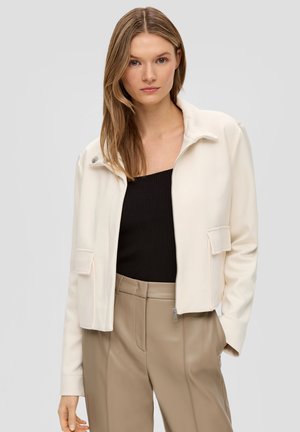 Crème gekorte jas met een ritssluiting aan de voorkant, zijzakken en een opstaande kraag. Gladde stof en subtiele naaddetails, gedragen over een zwarte top.