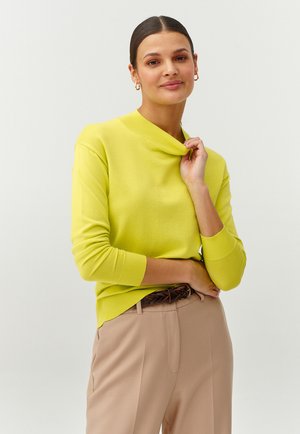 Pulover tricotat de un galben strălucitor, cu guler îndoitură și mâneci lungi, purtat cu pantaloni bej și un curea împletită maro.