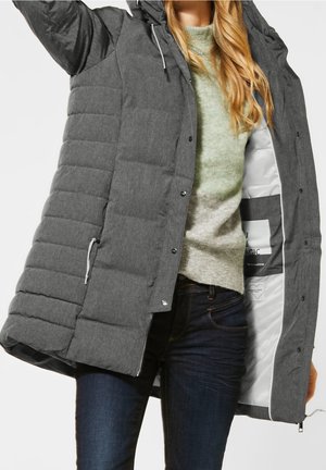 Femme portant un jean foncé et un pull vert clair ouvrant un long manteau d'hiver gris matelassé avec capuche, montrant la doublure intérieure blanche.