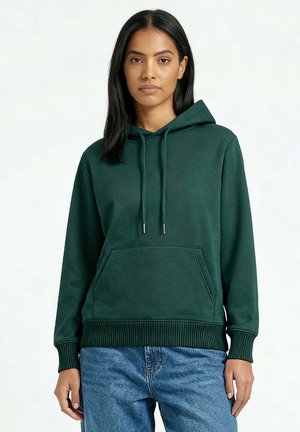 Donna che indossa una felpa verde scuro semplice con tasca frontale e jeans blu, in piedi davanti a uno sfondo grigio chiaro.