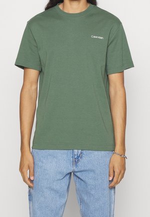 Camiseta verde de algodón con cuello redondo, mangas cortas y el logo bordado de Calvin Klein en el lado izquierdo del pecho. Se lleva con vaqueros azul claro.
