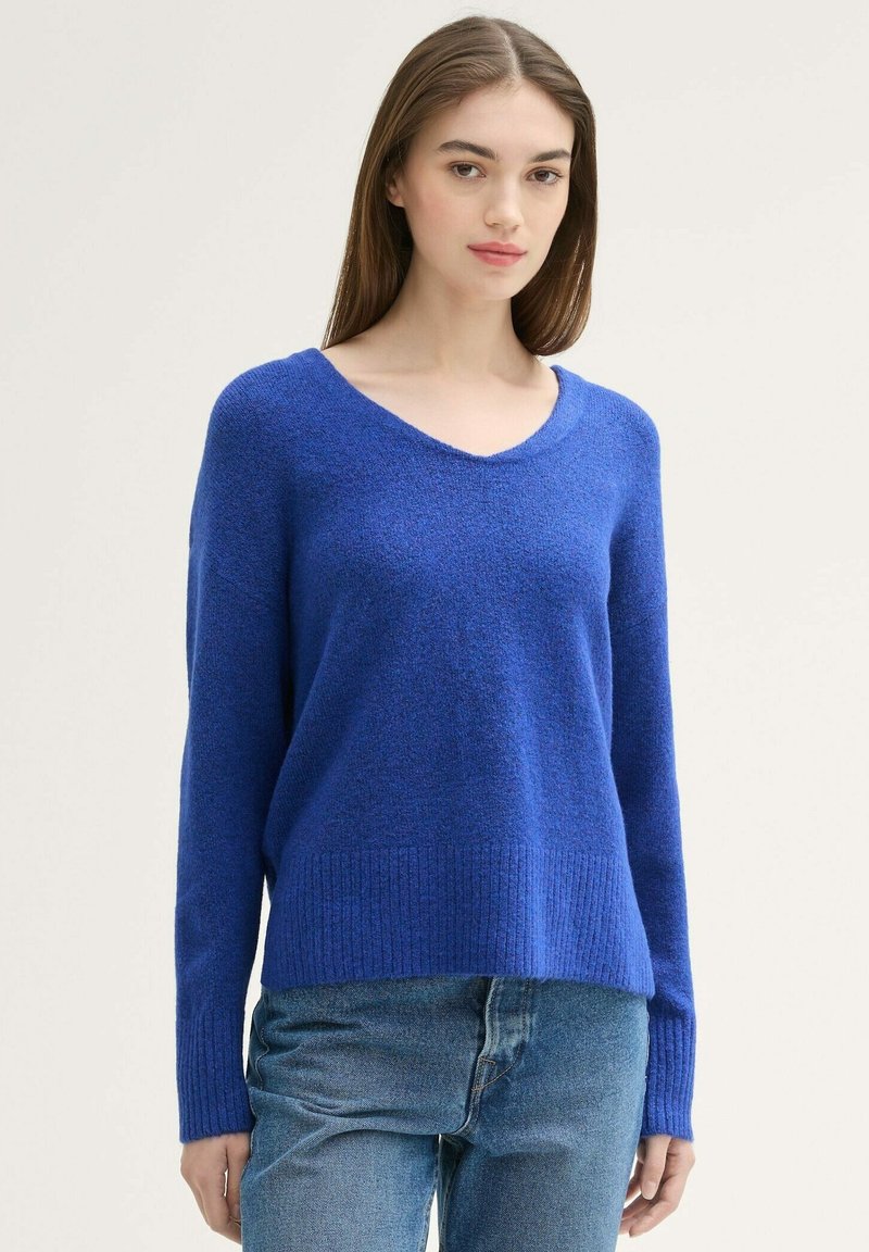 Blauwe gebreide trui met een v-hals, lange mouwen, geribbelde zoom en een textuurafwerking, gecombineerd met lichtblauwe jeans.