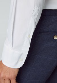 Weißes Langarmhemd mit Knopfmanschetten, kombiniert mit navyblauen, karierten Trousers. Der Stoff wirkt glatt mit einer knackigen Textur.