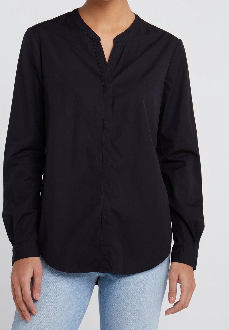 Femme portant un chemisier noir à manches longues avec un petit col en V et un jean bleu clair, debout devant un fond uni.