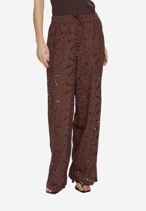 Pantalon marron à jambes larges avec motifs découpés complexes, taille élastique avec cordon de serrage, porté avec des sandales noires à talons et un haut marron.