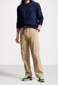 Marinblå sweatshirt med logga, beige löst sittande byxor med slitna detaljer och flerfärgade sneakers med varierande texturer och mönster.