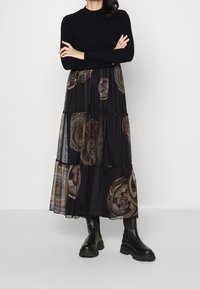 Gonna maxi nera a strati con motivi multicolori che si intrecciano, realizzata in tessuto trasparente con un design testurizzato. Indossata con stivaletti neri alla caviglia.