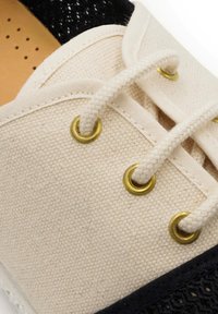 Canvas schoen met een beige bovenkant, zwarte gaasaccenten, gouden veterogen en witte veters. Textuur met zichtbare stikdetails.