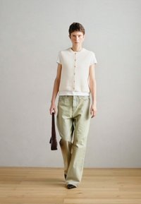 Λευκό ραβδωτό γιλέκο με κουμπιά συνδυασμένο με ανοιχτοπράσινα παντελόνια φαρδιάς γραμμής. Το άτομο κρατά μια σκούρα καφέ τσάντα, με ένα μινιμαλιστικό φόντο.