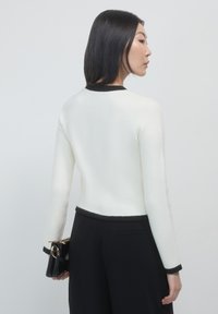 Weißer Strickpullover mit schwarzem Abschluss am Ausschnitt und Saum, lange Ärmel und schmaler Schnitt, kombiniert mit schwarzen Hosen.