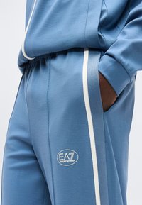 Μπλε αθλητικό παντελόνι με λευκή ρίγα στο πλάι και λογότυπο EA7 Emporio Armani κοντά στο ισχίο, χέρι ακουμπισμένο στην τσέπη.