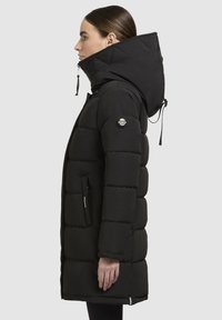 Sieviete profila skatījumā, ģērbusies garā melnā polsterētajā mētelī ar augstu apkakli un lielu kapuci, rokas atslābinātas pie sāniem uz vienkārša fona.