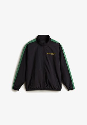 Chaqueta cortavientos negra con rayas laterales de cuadros verdes, diseño con cremallera, cuello alto y puños elásticos. Presenta detalles del logo en amarillo.