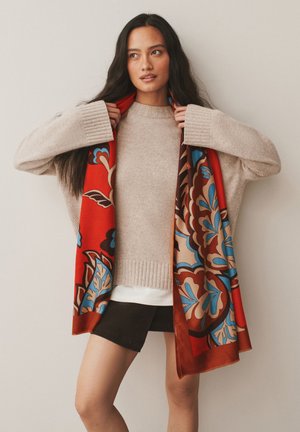 Femme en pull beige et short noir ajustant une écharpe rouge avec des motifs floraux bleus et marron sur un fond uni.