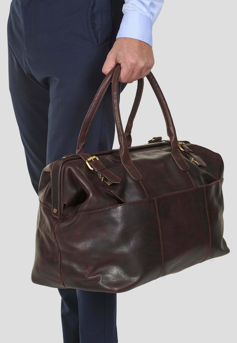 Brun lær duffelbag med doble hender, gulldetaljer og en strukturert overflate. Den har en strukturert form og synlige sømmer.