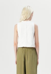 Witte mouwloze top met een cropped ontwerp, voorzien van een gestructureerde achternaad en rafelige randen, gecombineerd met losse olijfgroene broek.