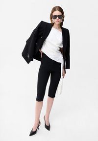 Frau trägt schwarze Cropped-Hose, weißes asymmetrisches Top, schwarzen Blazer lässig über die Schultern gelegt, schwarze High Heels und Sonnenbrille vor weißem Hintergrund.