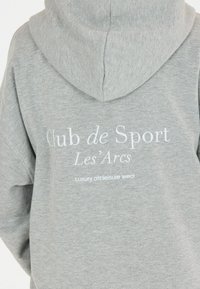 Grå hoodie med dragsko på huvan. Har vit text: "Club de Sport Les'Arcs" och "Luxury athleisure wear" tryckt på baksidan. Mjuk textur.