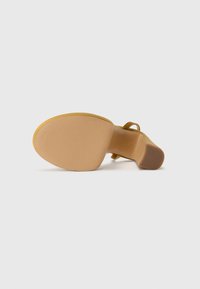 Scarpa con tacchi alti gialla, con tomaia liscia, retro aperto e sottile cinturino alla caviglia. La suola è color beige con una leggera texture e una forma arrotondata.