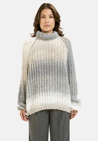 Frau trägt einen übergroßen, gestrickten Rollkragenpullover mit einem verlaufenden Grau-zu-Weiß-Muster und lockeren grauen Hosen und steht vor einem schlichten Hintergrund.