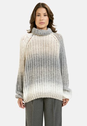 Frau trägt einen übergroßen, gestrickten Rollkragenpullover mit einem verlaufenden Grau-zu-Weiß-Muster und lockeren grauen Hosen und steht vor einem schlichten Hintergrund.