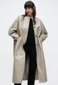 Žena s tmavými vlasmi má na sebe dlhý béžový kožený kabát cez čierny sveter a čierne nohavice, stojaca proti jednoliatemu bielemu pozadiu.